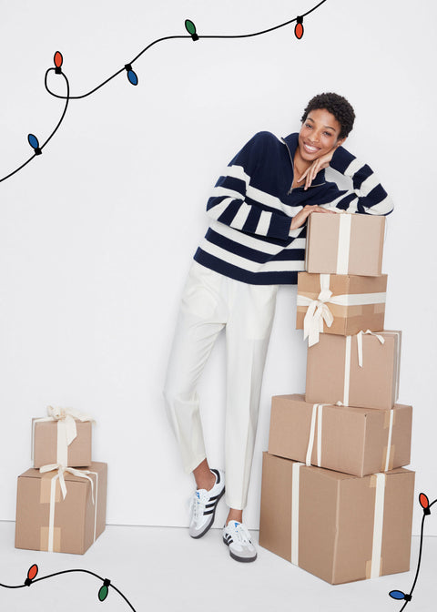 Model wearing Morgan sweater in navy/cream with Francis pant in cream leaning on stack of cardboard boxes wrapped in white bows.