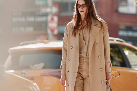 Female model wearing Rox trench in khaki with the Blanca shirt and City pants in sand walking by a taxi on the street
