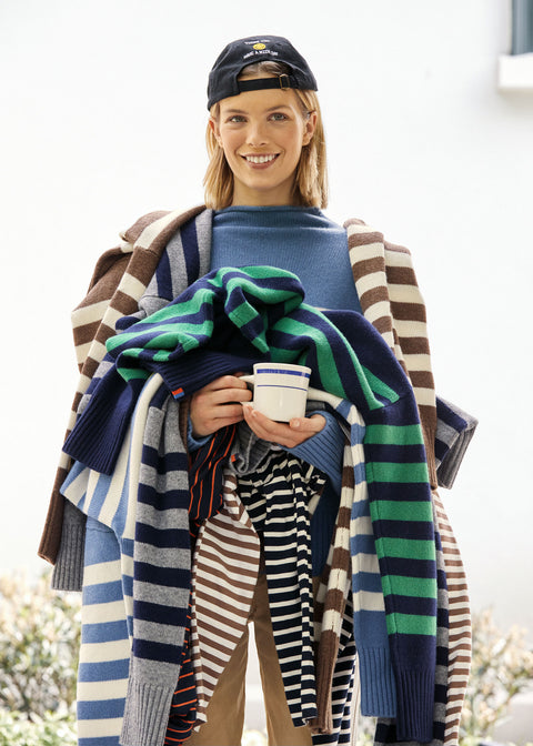 Model wearing the take out kap in navy backwards and is holding a pile of striped sweaters and tees